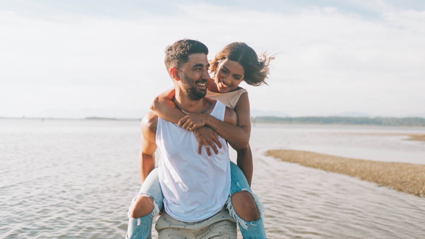 A smiling couple on the beach, one carrying the other piggyback